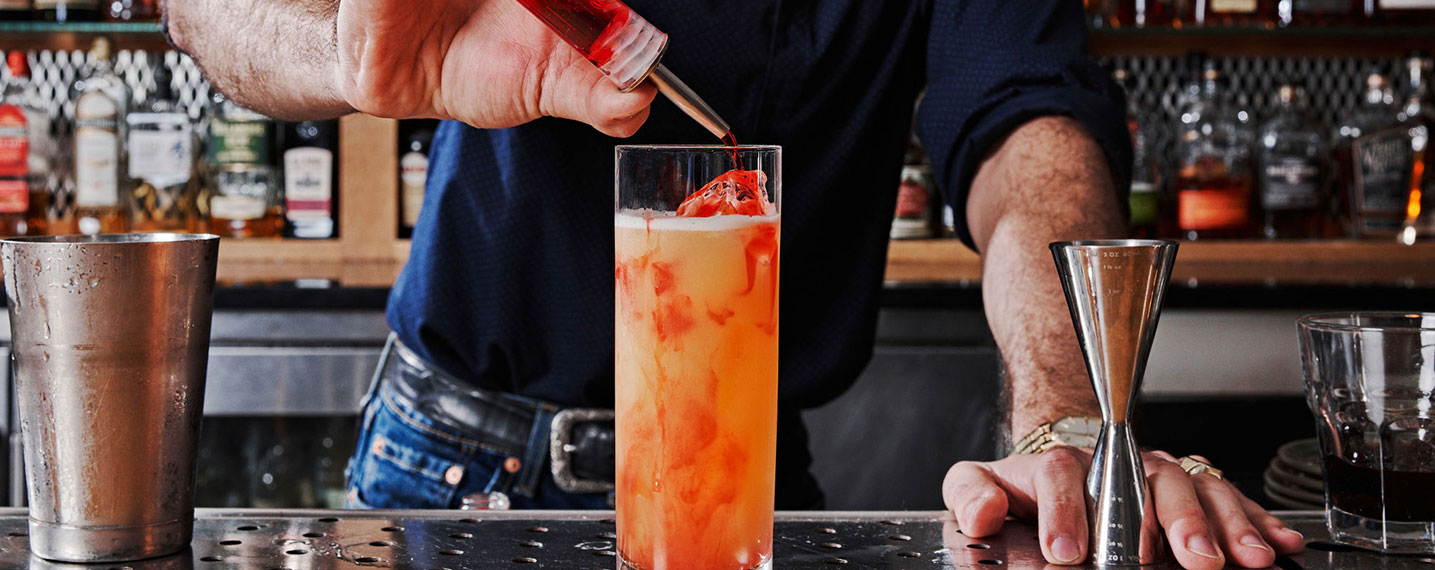 A bartender in a blue shirt pours red liquid from a bottle into a tall, orange cocktail glass on a wooden bar