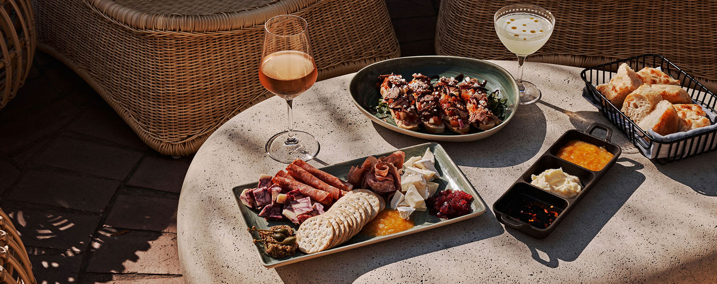 A stone patio table laden with appetizers, wine, and a cocktail, next to wicker chairs in sunlight